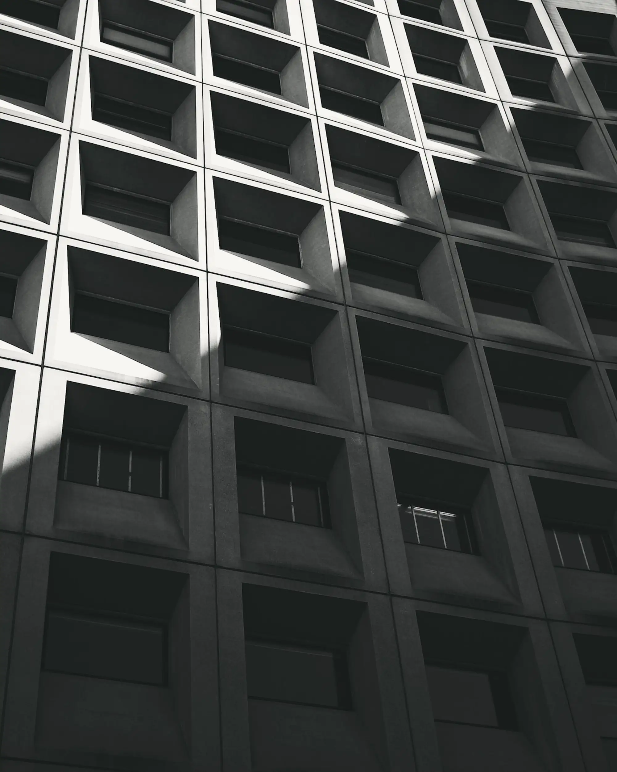 Fachada de celdas cuadradas en contrapicado, cortada en diagonal por una franja de luz de sol.